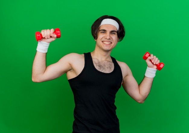 Jeune homme sportif portant des vêtements de sport et un bandeau travaillant avec des haltères souriant joyeusement