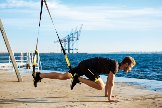 Jeune homme sportif s'entraînant avec trx près de la mer le matin.