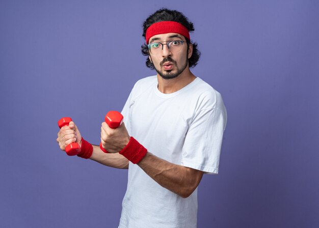 Jeune homme sportif surpris portant un bandeau avec un bracelet faisant de l'exercice avec des haltères