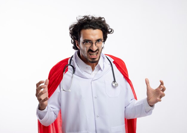 Jeune homme de super-héros caucasien agacé dans des lunettes optiques portant un uniforme de médecin avec une cape rouge et avec un stéthoscope autour du cou regardant la caméra