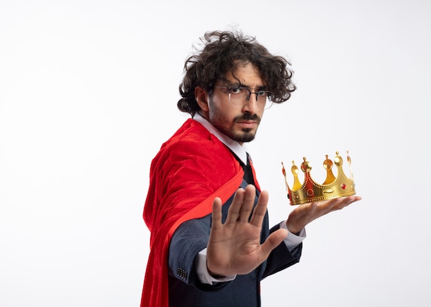 Un jeune homme de super-héros caucasien confiant dans des lunettes optiques portant un costume avec une cape rouge tient la couronne et les gestes arrêtent le signe de la main