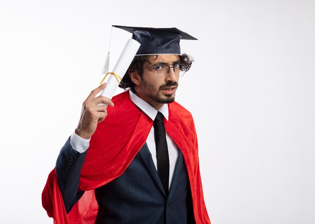 Un jeune homme de super-héros caucasien désemparé dans des lunettes optiques portant un costume avec une cape rouge et une casquette de graduation détient un diplôme