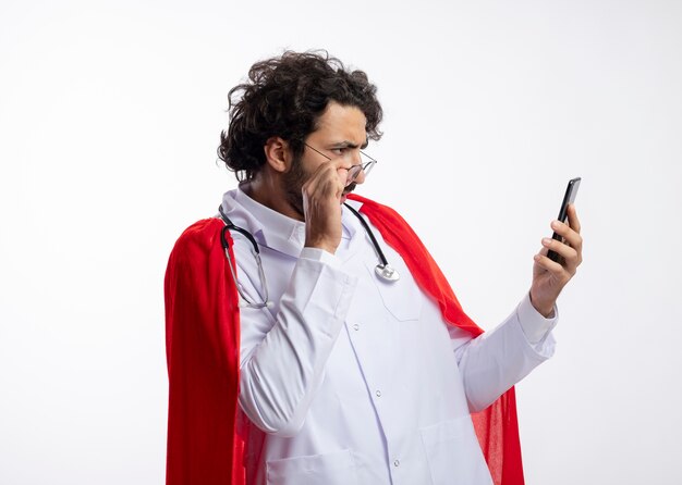 Jeune homme de super-héros caucasien ignorant dans des lunettes optiques portant un uniforme de médecin avec une cape rouge et avec un stéthoscope autour du cou tient et regarde le téléphone