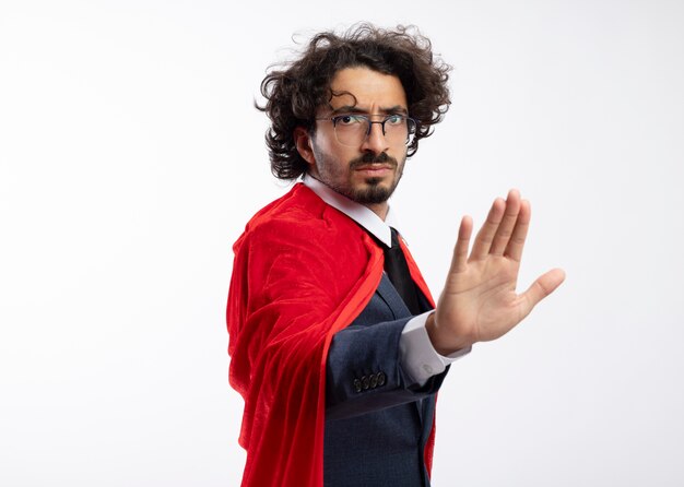 Jeune homme de super-héros caucasien sérieux dans des lunettes optiques portant un costume avec une cape rouge se tient sur le côté en gesticulant le signe de la main d'arrêt