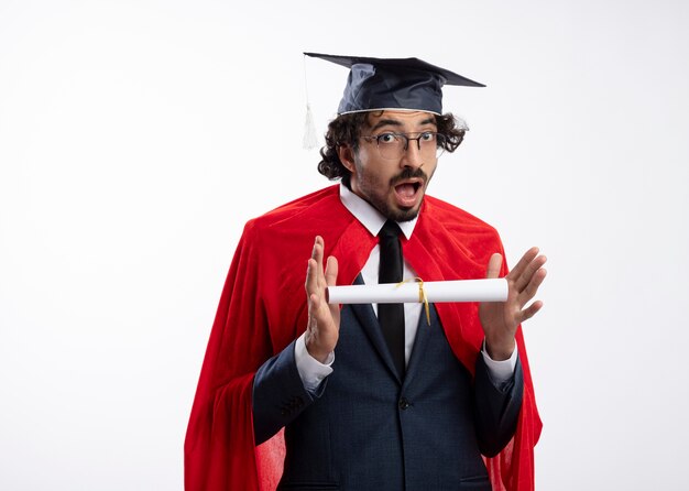 Photo gratuite un jeune homme de super-héros caucasien surpris en lunettes optiques portant un costume avec une cape rouge et une casquette de graduation détient un diplôme et regarde la caméra