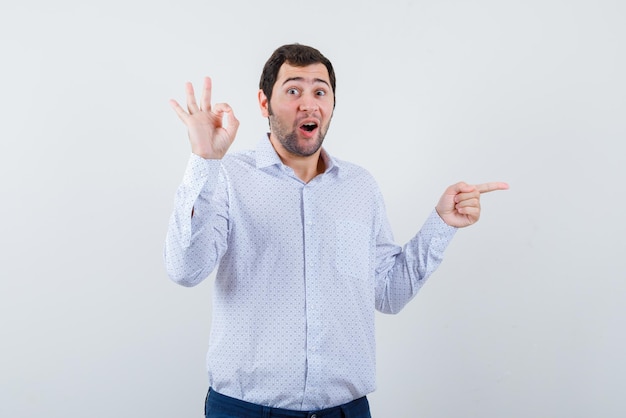 Jeune homme surprenant montrant un bon geste de la main et pointant quelque chose avec les doigts à droite sur fond blanc