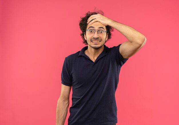 Jeune homme surpris en chemise noire avec des lunettes optiques tient la tête et regarde isolé sur un mur rose