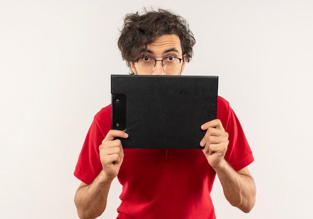 Jeune homme surpris en chemise rouge avec des lunettes optiques détient le presse-papiers et semble isolé sur un mur blanc