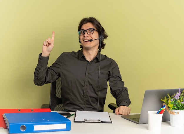 Jeune homme surpris employé de bureau sur les écouteurs dans des lunettes optiques est assis au bureau avec des outils de bureau à l'aide d'un ordinateur portable pointe vers le haut à côté isolé sur fond vert avec espace de copie