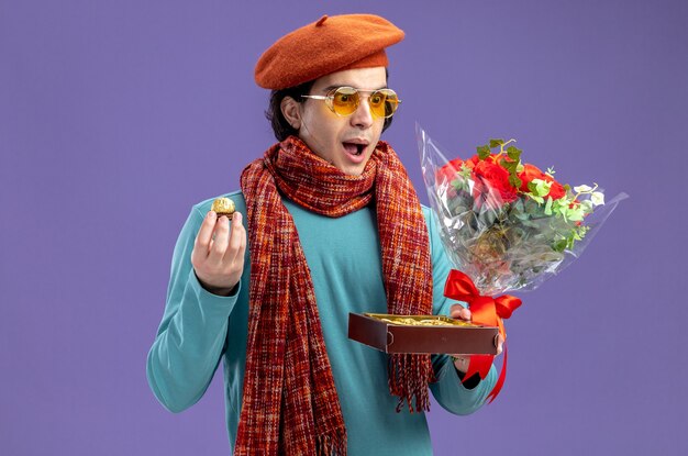 Jeune homme surpris le jour de la Saint-Valentin portant un chapeau avec une écharpe et des lunettes tenant et regardant le bouquet avec une boîte de bonbons isolés sur fond bleu