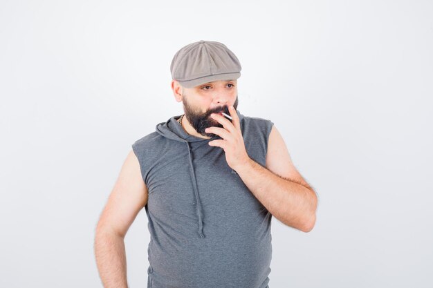 Jeune homme en sweat à capuche sans manches, casquette fumant tout en posant et en ayant l'air confiant, vue de face.