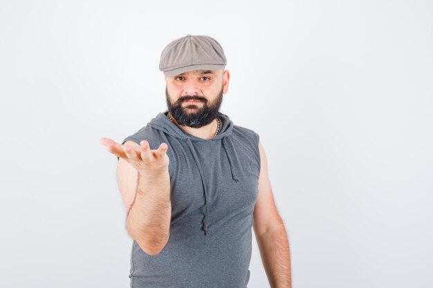 Jeune homme en sweat à capuche sans manches, casquette levant la main dans une pose de questionnement et l'air confiant, vue de face.