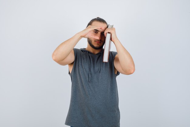 Jeune homme en sweat à capuche sans manches tenant un livre sur l'épaule tout en montrant le signe de la taille et à la grave, vue de face.