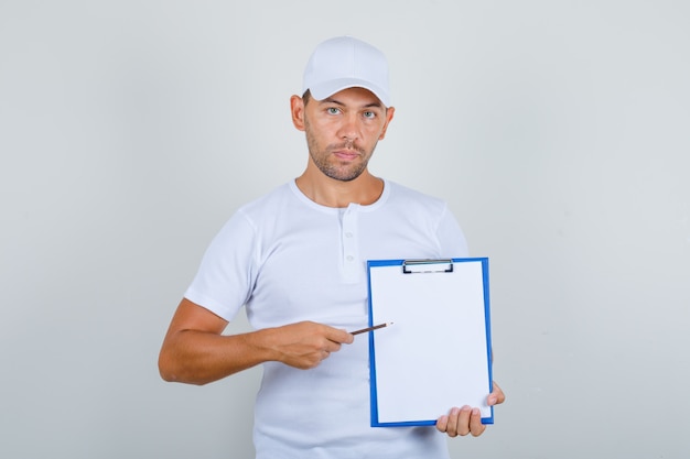 Jeune homme en t-shirt blanc et casquette montrant des notes sur le presse-papiers avec un crayon, vue de face.