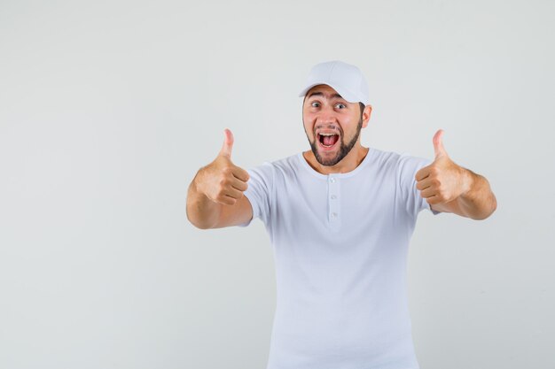 Jeune homme en t-shirt blanc, casquette montrant le pouce vers le haut tout en souriant et en regardant joyeux, vue de face.