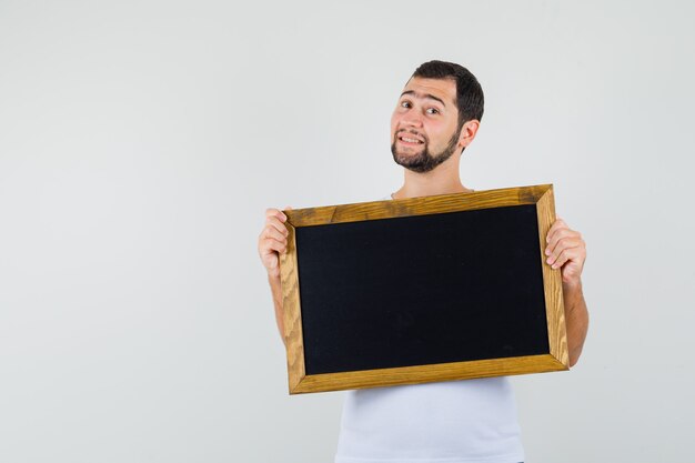 Jeune homme en t-shirt blanc debout avec tenant un tableau noir et à la vue de face, prêt.