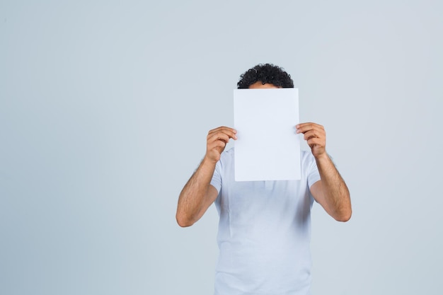 Jeune homme en t-shirt blanc gardant une feuille de papier vierge et ayant l'air confiant, vue de face.