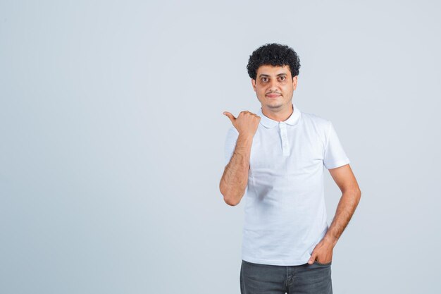 Jeune homme en t-shirt blanc et jeans mettant la main dans la poche tout en pointant du doigt et en ayant l'air sérieux, vue de face.
