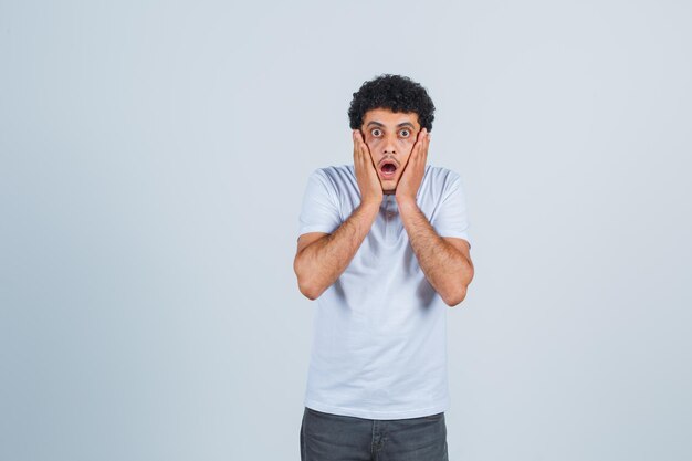 Jeune homme en t-shirt blanc et jeans mettant les mains sur les joues, ouvrant la bouche et l'air surpris, vue de face.