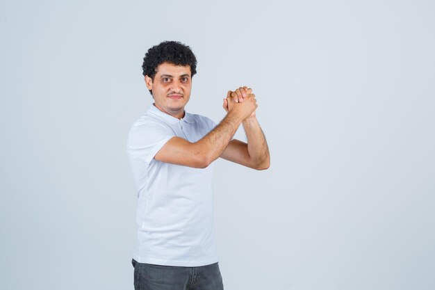 Jeune homme en t-shirt blanc et jeans serrant les mains et l'air heureux, vue de face.