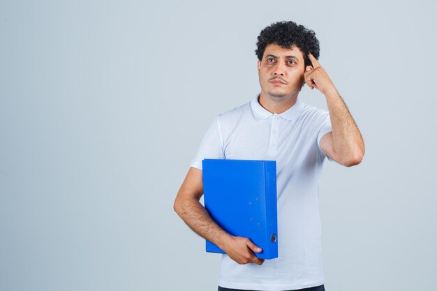 Jeune homme en t-shirt blanc et jeans tenant un dossier et mettant le doigt sur la tempe et l'air pensif, vue de face.