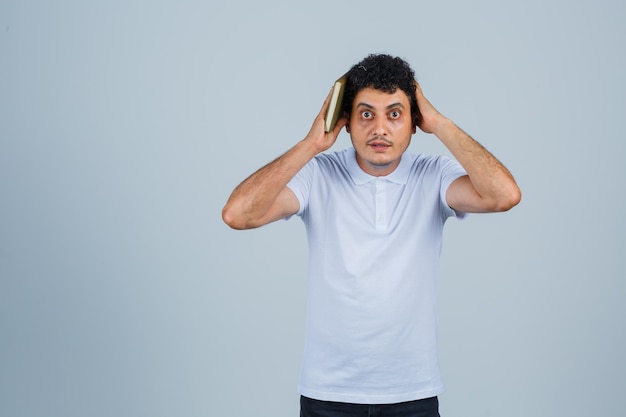 Jeune homme en t-shirt blanc et jeans tenant un livre et appuyant sur les oreilles et l'air surpris, vue de face.