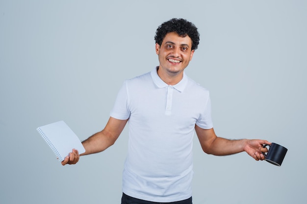 Jeune homme en t-shirt blanc et jeans tenant une tasse de thé tout en montrant un ordinateur portable et l'air heureux, vue de face.