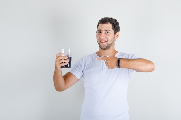 Jeune homme en t-shirt blanc montrant un verre de cola avec le doigt et l'air heureux