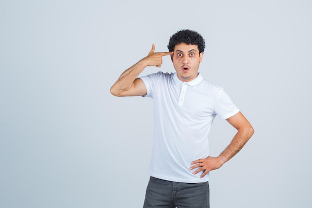 Jeune homme en t-shirt blanc, pantalon faisant un geste de suicide et l'air surpris, vue de face.