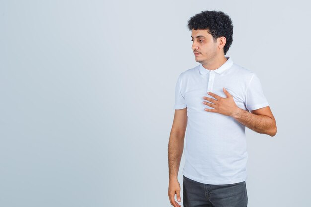 Jeune homme en t-shirt blanc, pantalon gardant la main sur la poitrine et l'air pensif, vue de face.