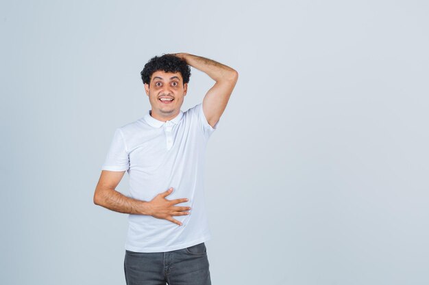 Jeune homme en t-shirt blanc, pantalon gardant la main sur la tête et l'air étonné, vue de face.