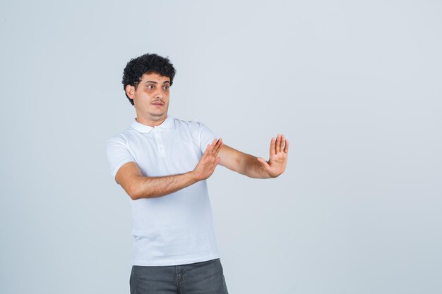 Jeune homme en t-shirt blanc, pantalon montrant un geste d'arrêt et ayant l'air effrayé, vue de face.