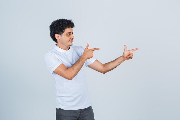 Jeune homme en t-shirt blanc, pantalon montrant le geste du pistolet et l'air confiant, vue de face.