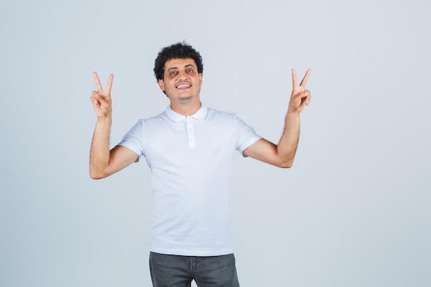 Jeune homme en t-shirt blanc, pantalon montrant le geste de la victoire et l'air confiant, vue de face.