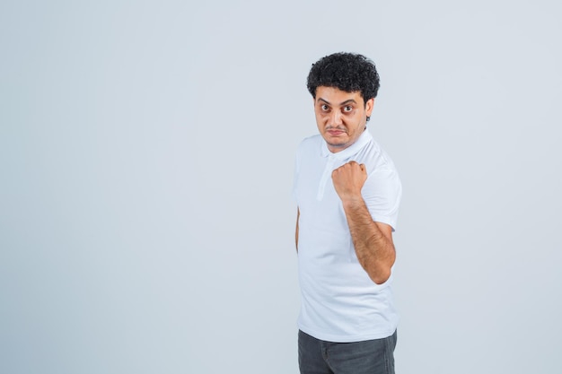 Jeune homme en t-shirt blanc, pantalon montrant le poing levé et l'air confiant, vue de face.