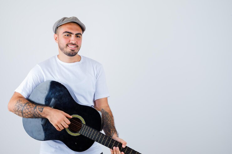 Photo gratuit jeune homme en t-shirt blanc, pantalon noir, casquette grise jouant de la guitare et ayant l'air heureux