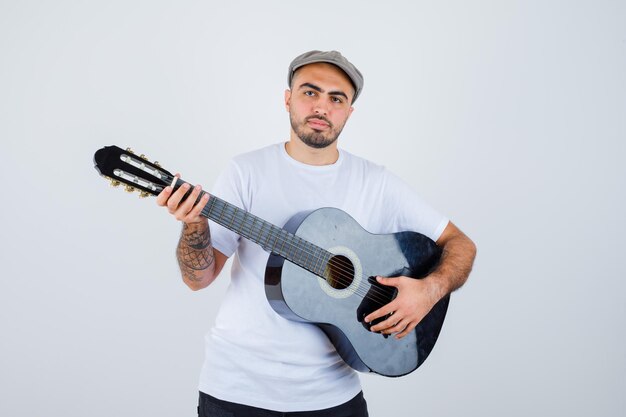 Jeune homme en t-shirt blanc, pantalon noir, casquette grise jouant de la guitare et ayant l'air sérieux