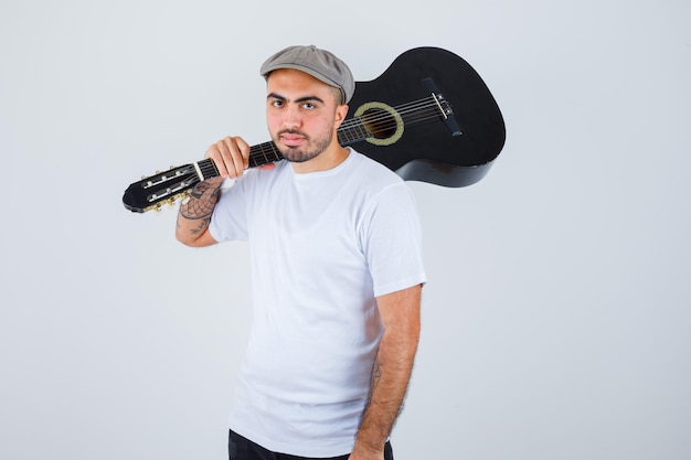 Jeune homme en t-shirt blanc, pantalon noir, casquette grise tenant une guitare et ayant l'air sérieux