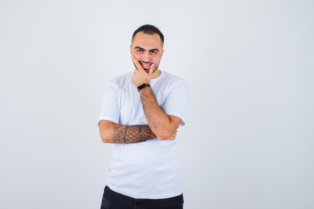 Jeune homme en t-shirt blanc et pantalon noir mettant la main sur le menton et l'air heureux