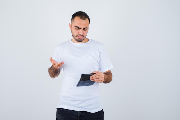 Jeune homme en t-shirt blanc et pantalon noir tenant une calculatrice et s'étirant la main de manière interrogative et l'air perplexe