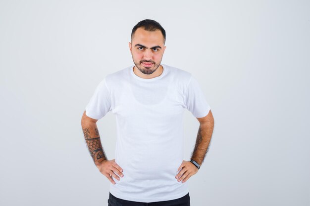 Jeune homme en t-shirt blanc et pantalon noir tenant les mains sur la taille et l'air heureux