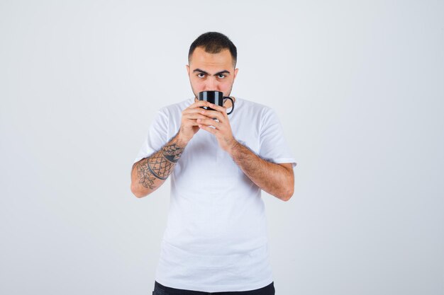 Jeune homme en t-shirt blanc et pantalon noir tenant une tasse de thé et la buvant et ayant l'air sérieux