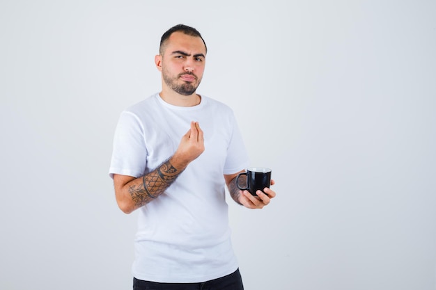 Jeune homme en t-shirt blanc et pantalon noir tenant une tasse de thé et montrant un geste italien et l'air heureux