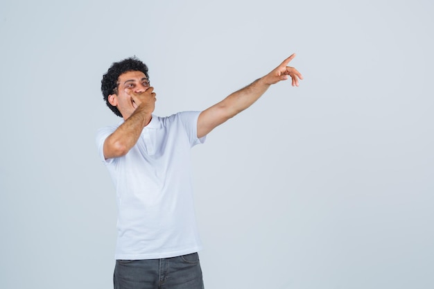 Jeune homme en t-shirt blanc, pantalon pointant vers quelque chose et ayant l'air heureux, vue de face.