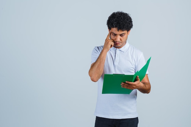 Jeune homme en t-shirt blanc, pantalon regardant par-dessus les notes dans le presse-papiers et ayant l'air pensif, vue de face.