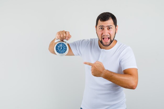 Jeune homme en t-shirt blanc pointant sur l'horloge et à la vue de face, impatient.