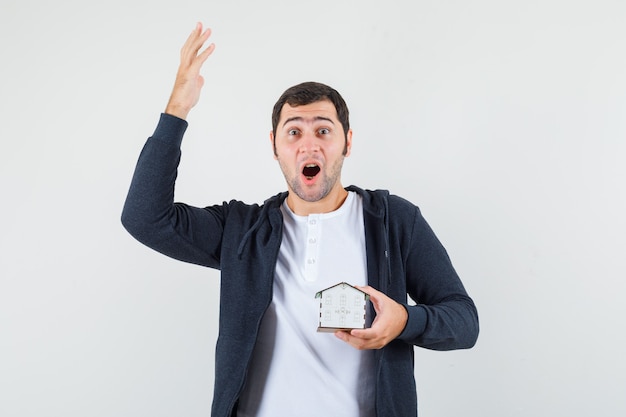 Jeune homme en t-shirt blanc et sweat à capuche noir à glissière avant tenant le modèle de la maison et levant la main en geste eureka et à la vue excitée, vue de face