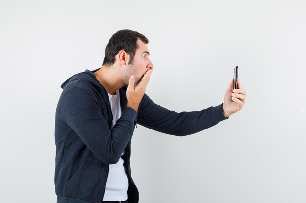 Jeune homme en t-shirt blanc et sweat à capuche noir à glissière devant parler à quelqu'un par vidéoconférence et à la surprise, vue de face.