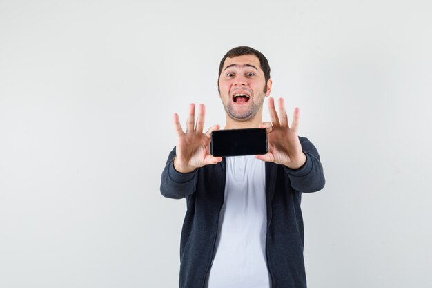 Jeune homme en t-shirt blanc et sweat à capuche noir zippé sur le devant tenant le smartphone à deux mains et à la vue de face, heureux.