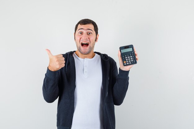 Jeune homme en t-shirt blanc et sweat à capuche noir zippé tenant la calculatrice et montrant le pouce vers le haut et à l'optimiste, vue de face.